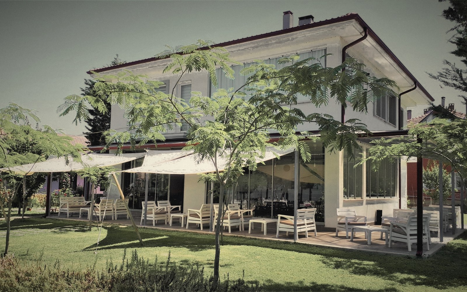 BEYAZ EV | Private Desk’in bahçesi: iki katlı beyaz yapı, zemin katta boydan boya cam yüzey, önünde gerilmiş beyaz gölge yelkenleri ve pergola, ahşap teras üzerinde beyaz bahçe koltukları, biçilmiş çim ve genç akasyalar.